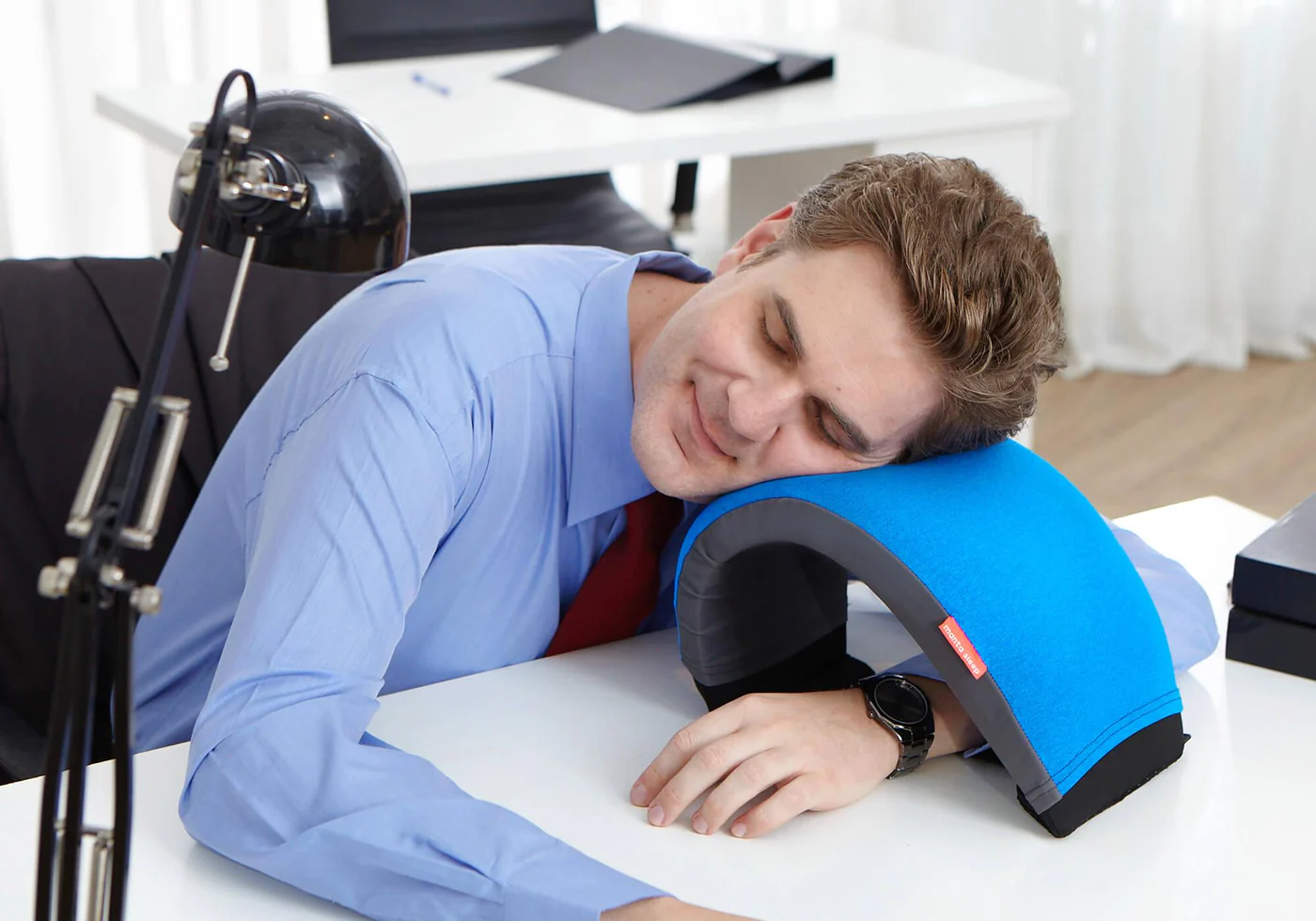 A man taking a coffee nap on his desk, using a blue nap pillow with an arc design.
