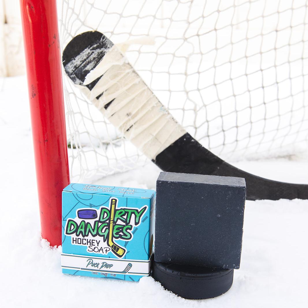 A black soap bar sits in the snow with a hockey goal, hockey puck and a hockey stick. Puck Drop