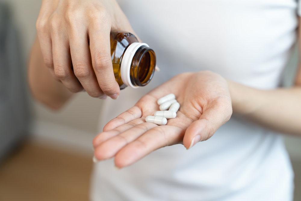 Close-up of a person in a white shirt pouring white capsules from a brown pill bottle into their open palm.