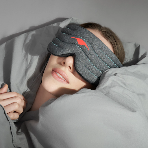 A girl wearing a weighted eye mask for insomnia lying down surrounded by gray pillows.