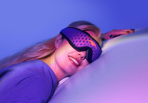 A smiling blonde girl wearing the Manta Sleep Mask PRO while lying down on a pillow