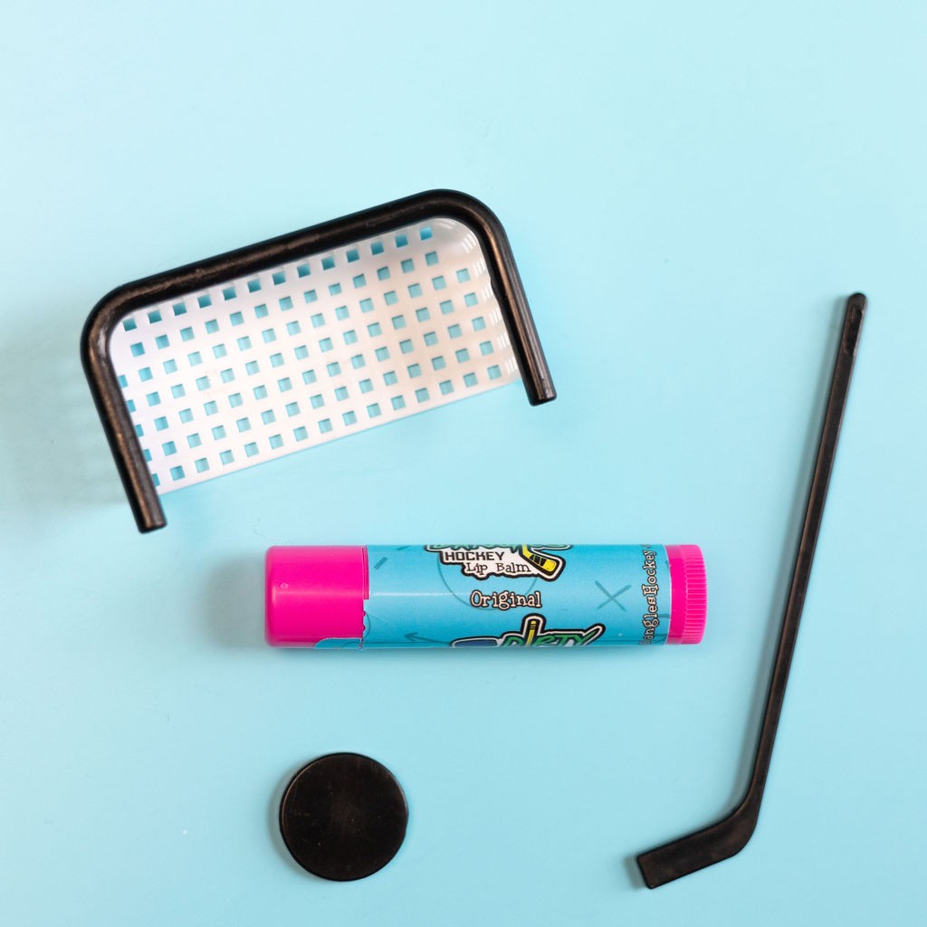 A pink stick of original flavor lip balm on a blue background with small hockey toys. Dirty dangles natural lip balm
