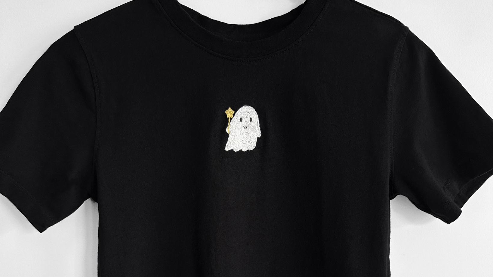A small white ghost holding a yellow star, embroidered on the chest of a black t-shirt.