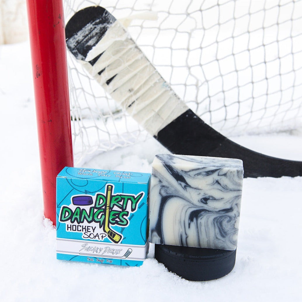 A black and white soap bar with a blue box. Sneaky deles scent. on a snowy background with a hockey net and hockey stick.