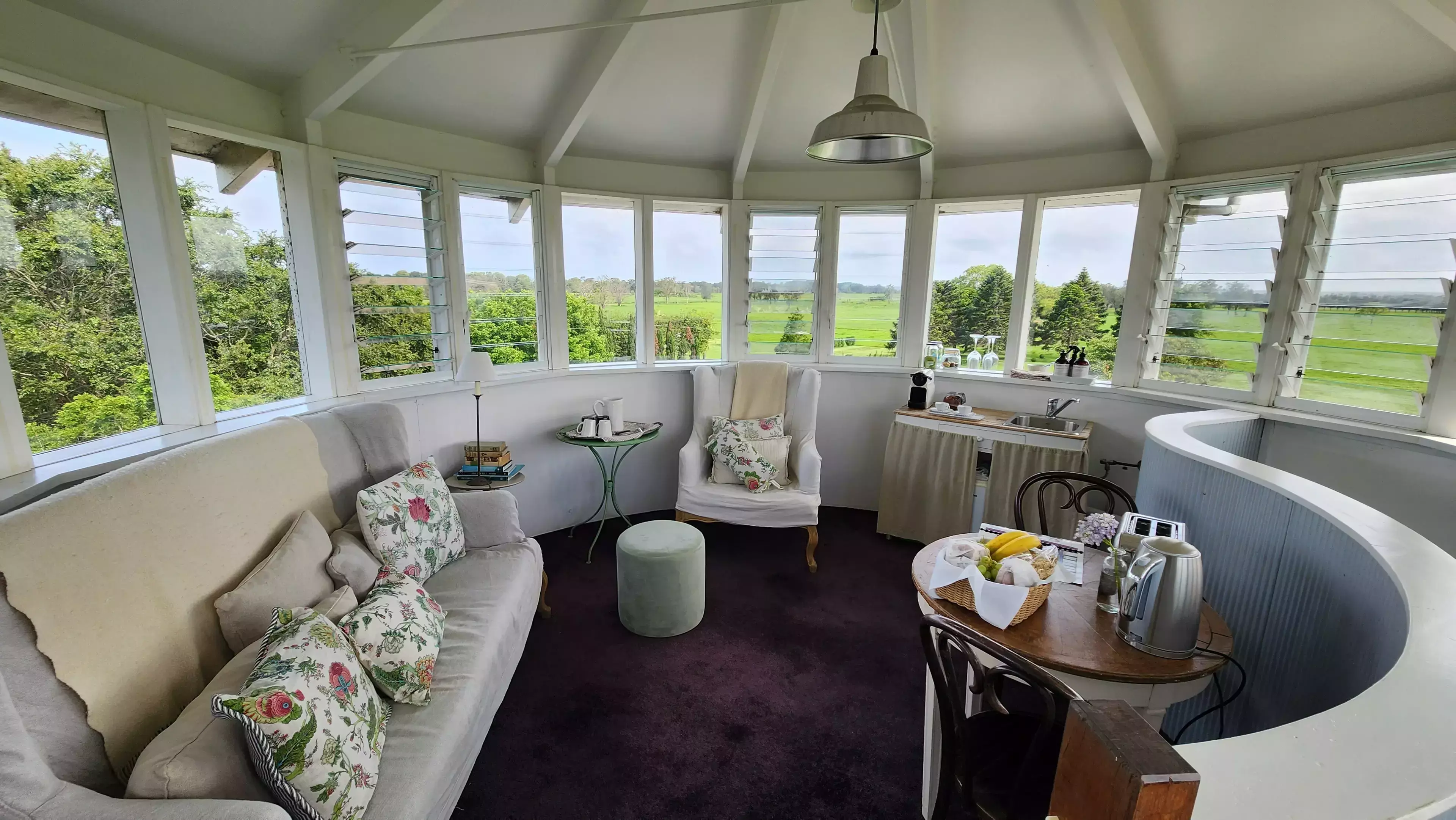 Light-filled circular sitting room inside a converted silo, featuring wraparound louvre windows with countryside views, a cosy sofa and armchair, small dining table with breakfast setup, compact kitchenette, and white timber ceiling beams.