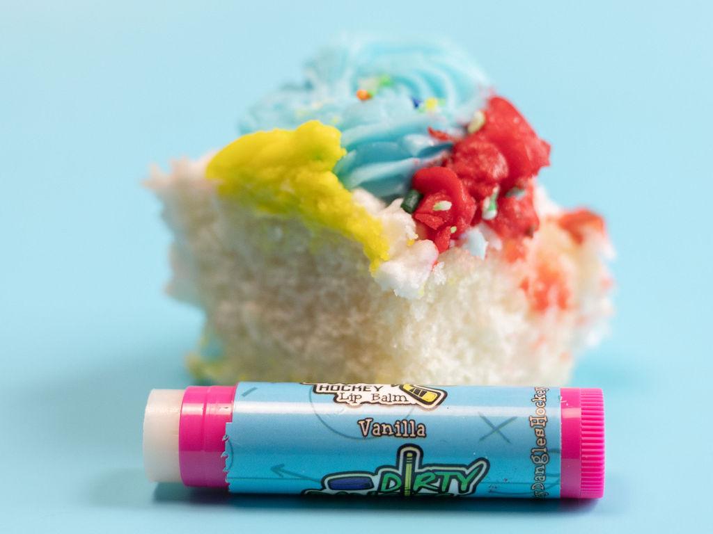 A pink stick of dirty dangles natural lip balm. Vanilla scent on a blue background with a piece of vanilla cake.