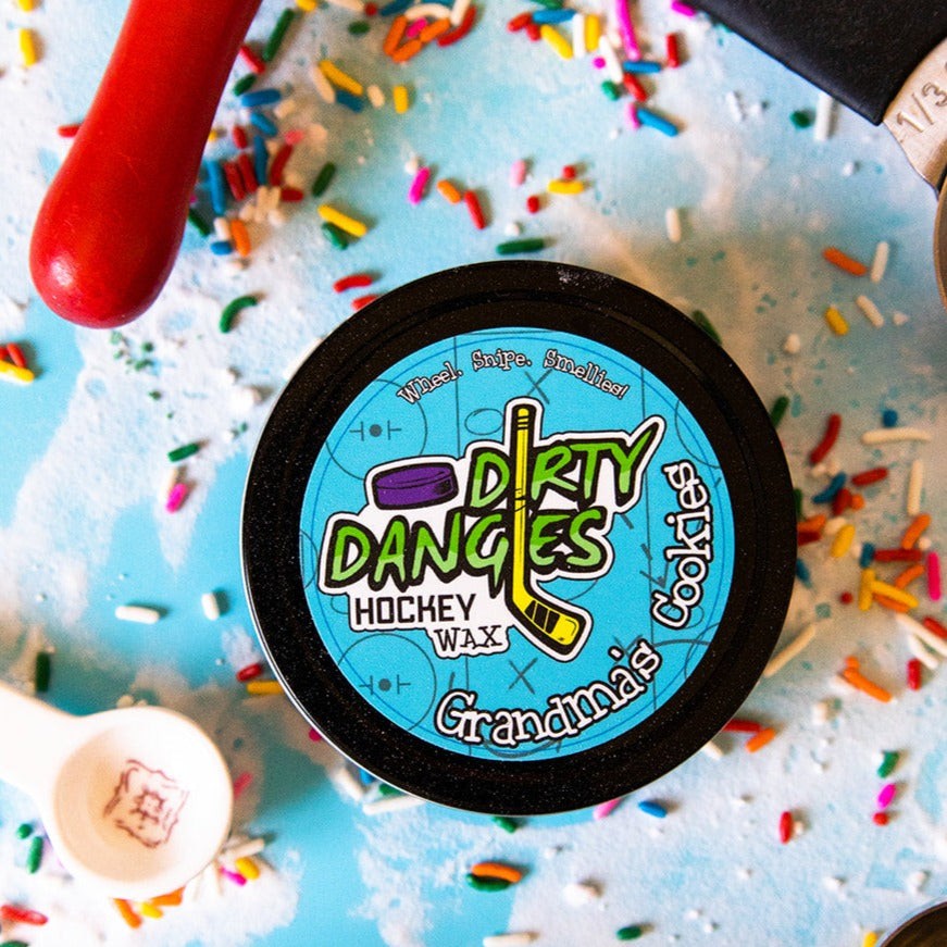 A tin of dirty dangles hockey stick wax grandma's cookies scent on a blue background with sugar, sprinkles and baking equipment.