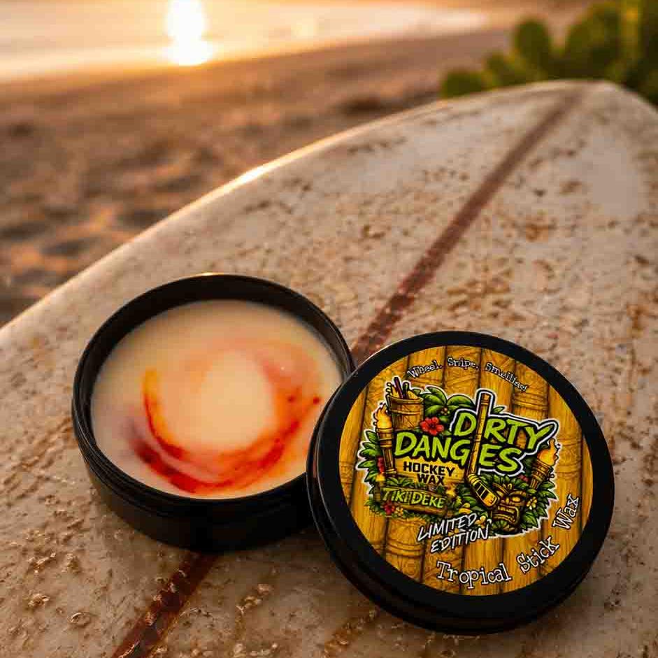 An open tin of dirty dangles tiki deke limited edition tropical stick wax on a rustic surfboard at the beach