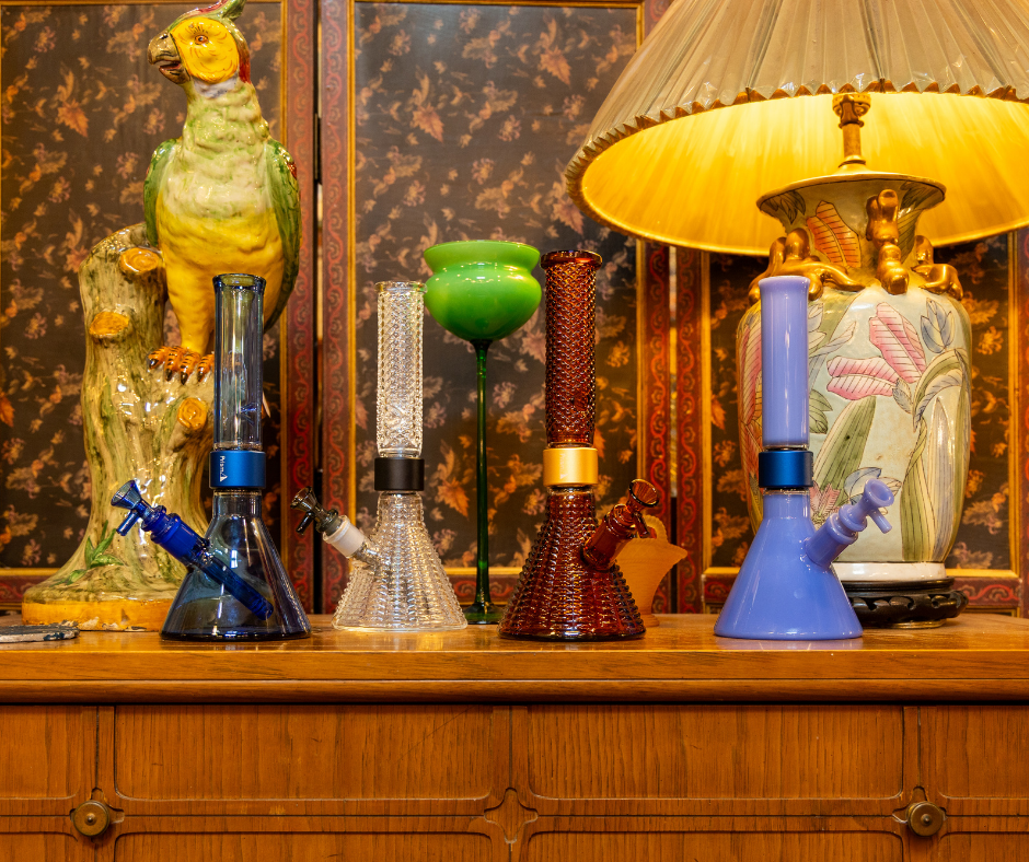 Four Prism MINI Beaker Bongs displayed on a mid-century modern desk alongside a clay parrot sculpture and antique lamp. The setup integrates the textural Jane West Art Deco clear and amber bongs with two MINI Monochrome Beakers in transparent sapphire and opaque blueberry, showcasing the full aesthetic range of the modular collection.
