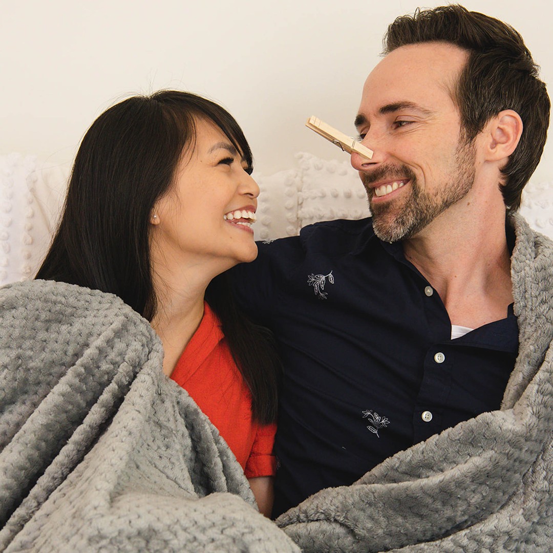 A smiling couple sits on a couch under a gray blanket. The man has a clothespin on his nose.