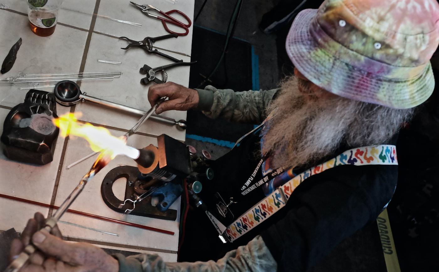 Pioneering glass artist Bob Snodgrass, the "Godfather of Glass," creating a pipe in his hot shop studio, surrounded by tools of the trade and a glass torch. The image highlights the detailed craftsmanship and artistic heritage that defines premium borosilicate glass pipes.