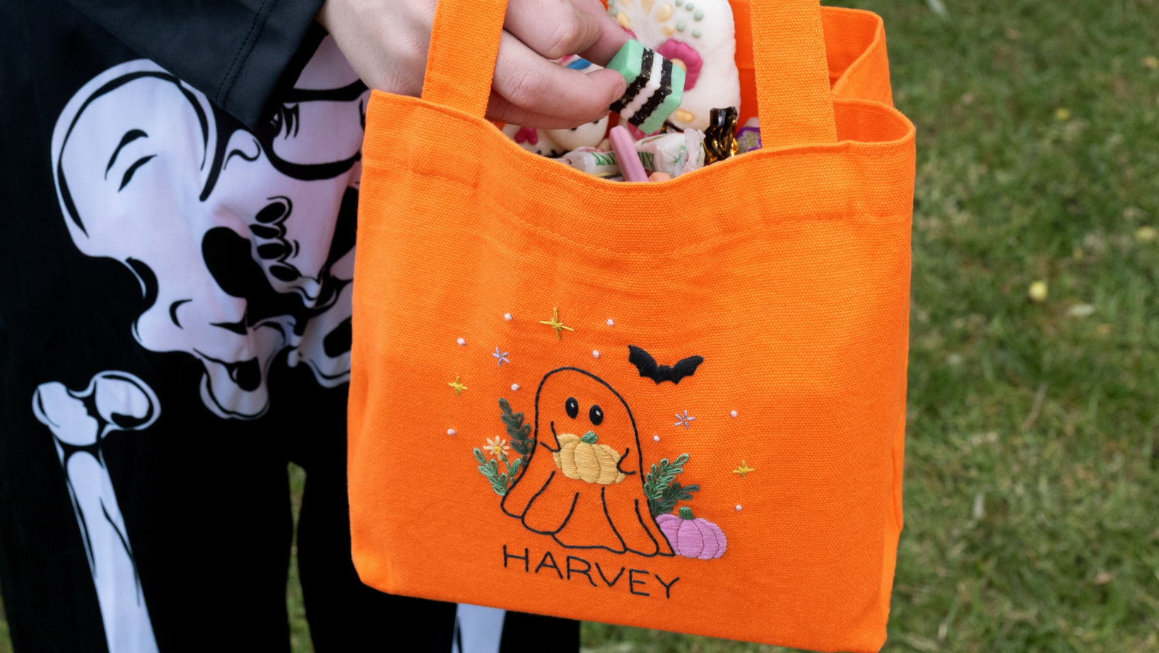 A child in a skeleton costume holding an orange trick-or-treat bag embroidered with a ghost and the name Harvey.