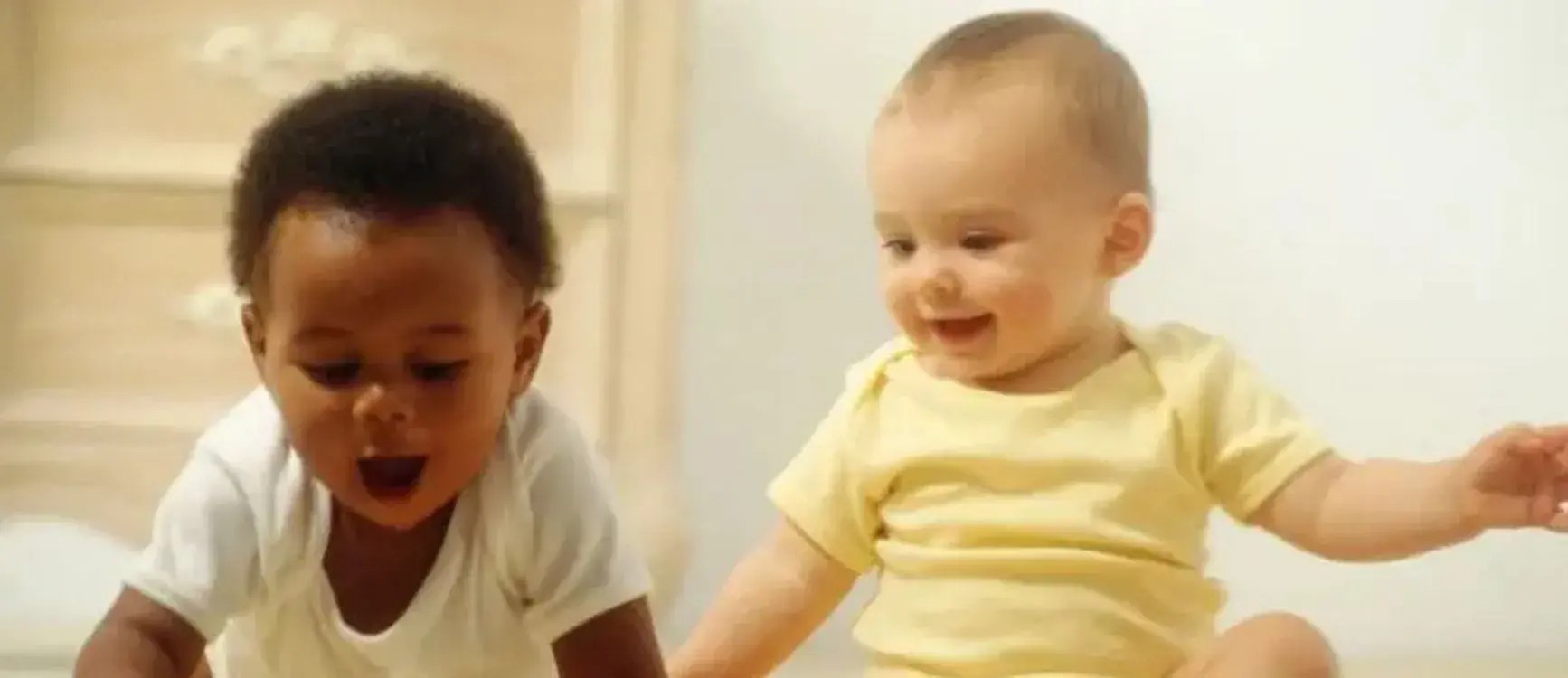 Two babies are happily playing together on a soft bed, surrounded by colorful toys and blankets.