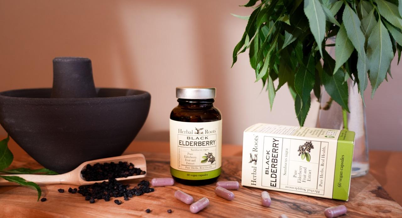 Bottle of Herbal Roots Black Elderberry next to a large mortar and pestle with a wooden spoon laying on the table filled with black elderberries. There are capsules on the table in front of the bottle.
