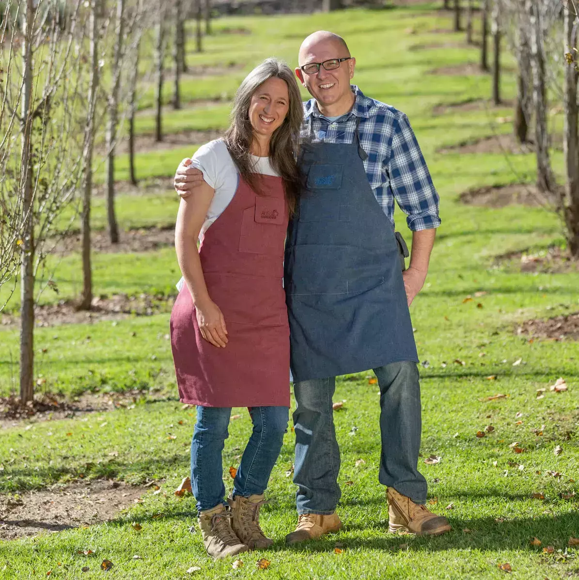 Brian and Kaylene wearing aprons stand arm in arm between rows of young trees in a sunlit orchard.
