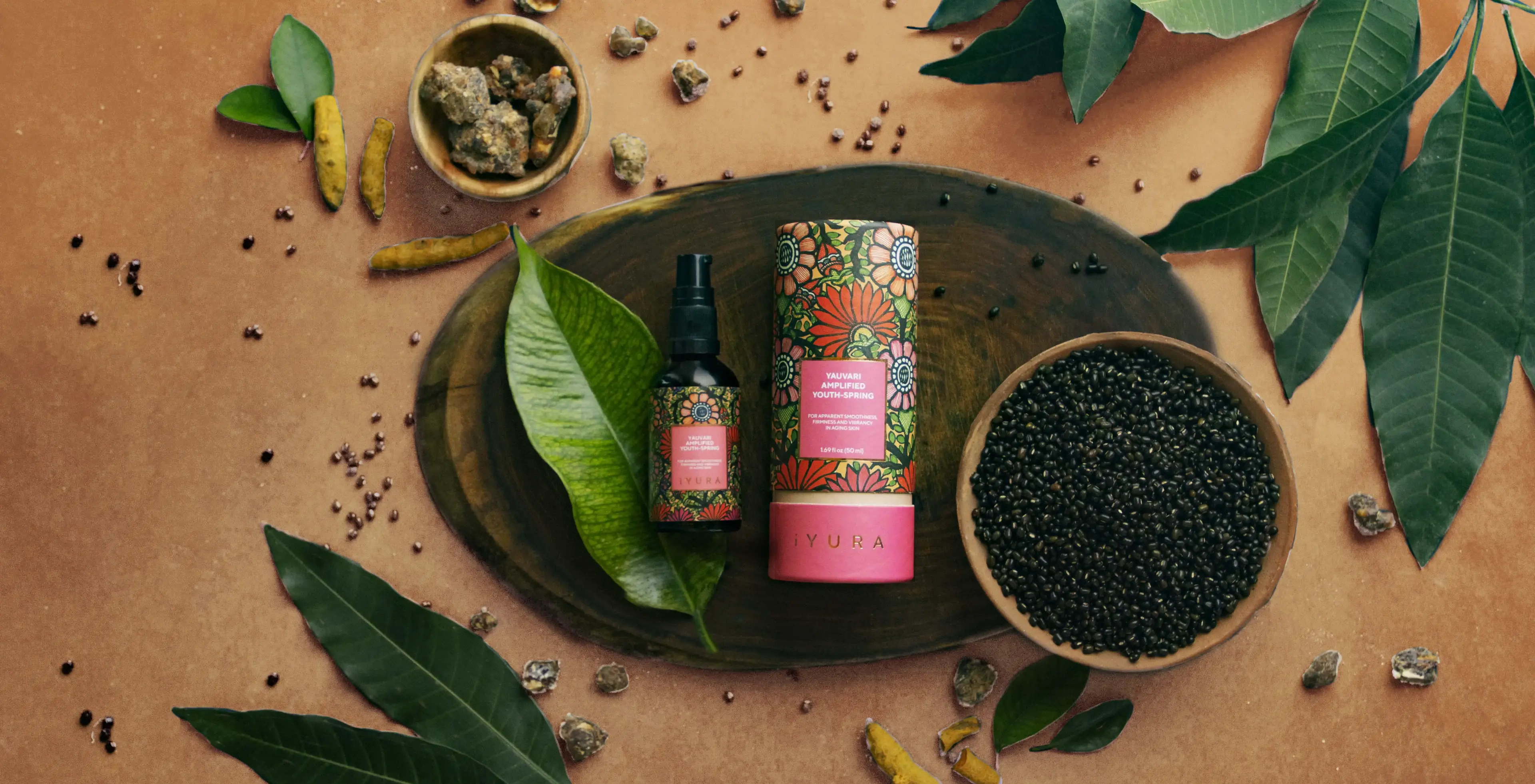 A bottle of Yauvari kept over a wooden plate, with a clay bowl filled with black gram, surrounded by mango leaves, turmeric and more over an earthy brown color background.