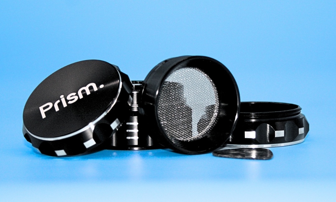 Disassembled black 4-piece Prism Herb Grinder showing the modular components: branded logo lid, diamond-cut grinding chamber, thick micron-mesh kief screen, and collection chamber with a branded KOZO guitar pick kief scraper.