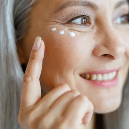 close-up of older woman applying 3 dots of eye cream underneath her eye.