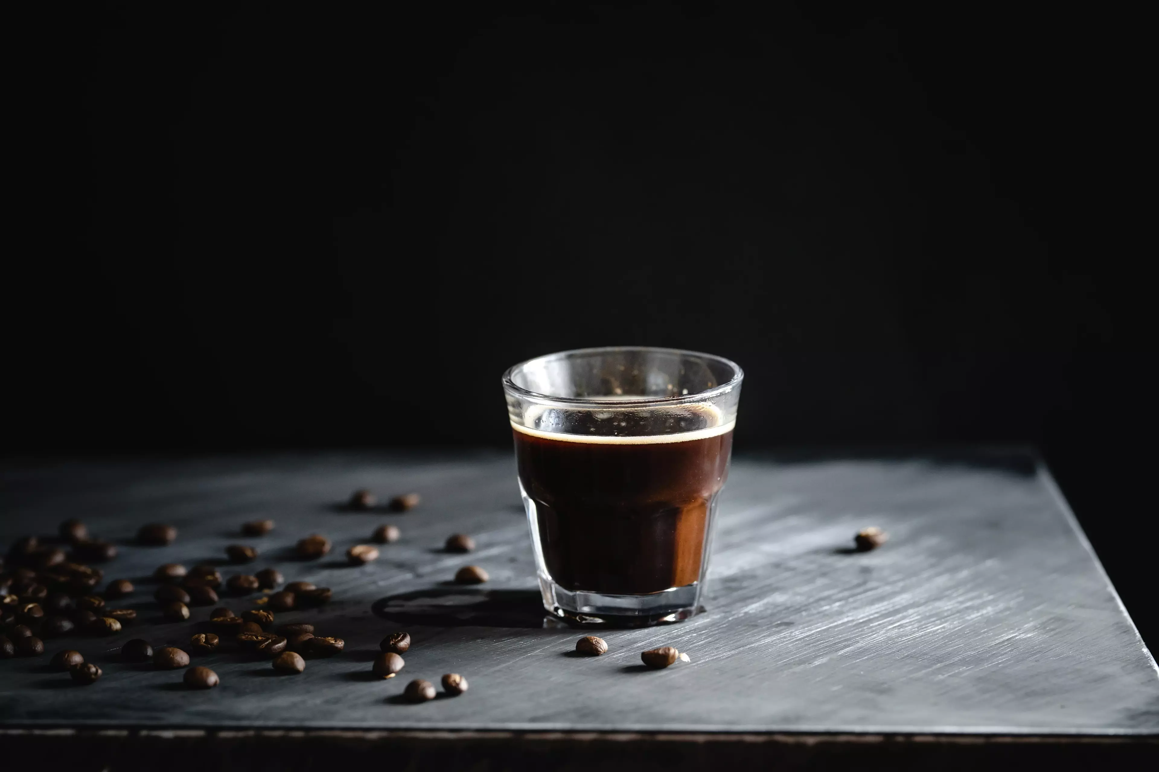 Fresh espresso shot in a clear glass on a dark background with scattered coffee beans.