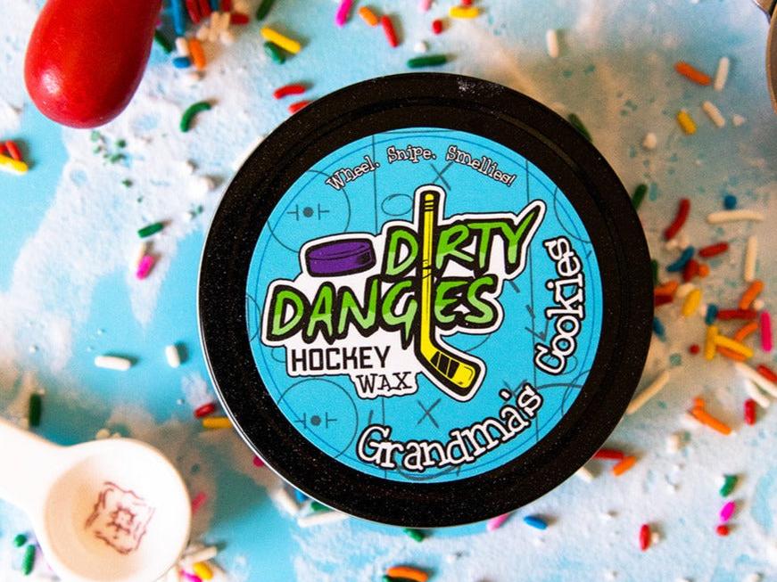 A tin of Dirty Dangles Hockey Stick Wax Grandma's Cookies Scent sits on a blue background surrounded by sprinkles and baking supplies