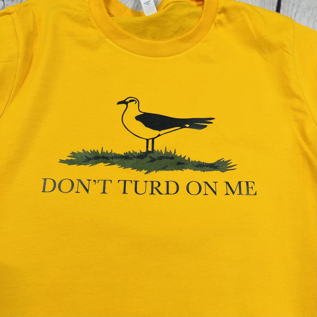 Close up of a gold and yellow t shirt with a picture of a seagull standing in the grass. Text on the shirt says "Don't Turd On Me." Similar to the Gadsden "Dont Tread On Me" Flag