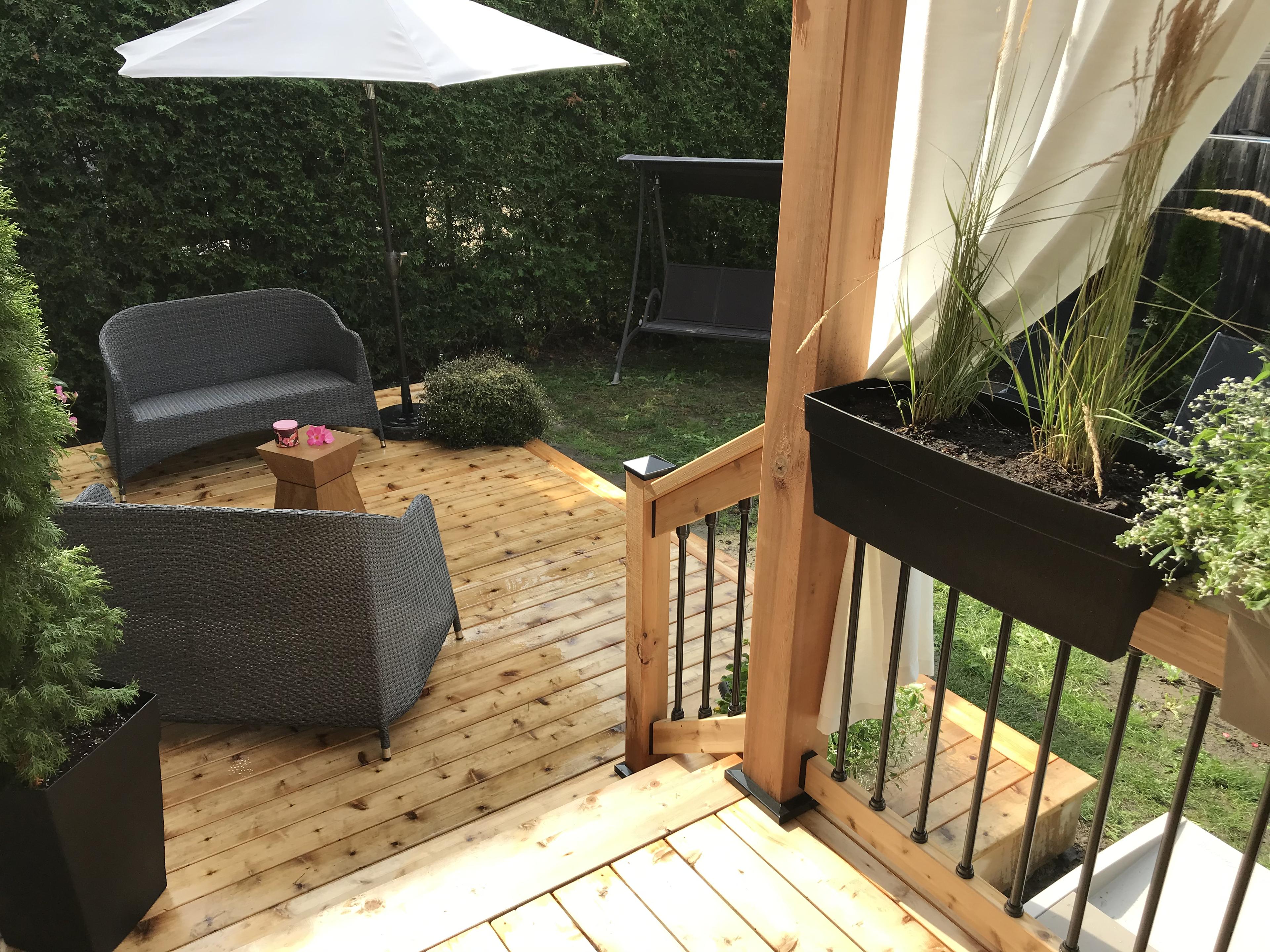 Image of a beautiful backyard with wooden stair case and deck. the railing is adorned with a white flowing curtain and the deck is decorated with seating and an umbrella