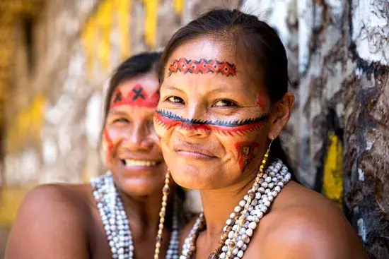 Two Indigenous women from Amazon smiling, wearing traditional face paint in red, black, and white patterns, along with layered bead necklaces, standing against a painted stone wall.