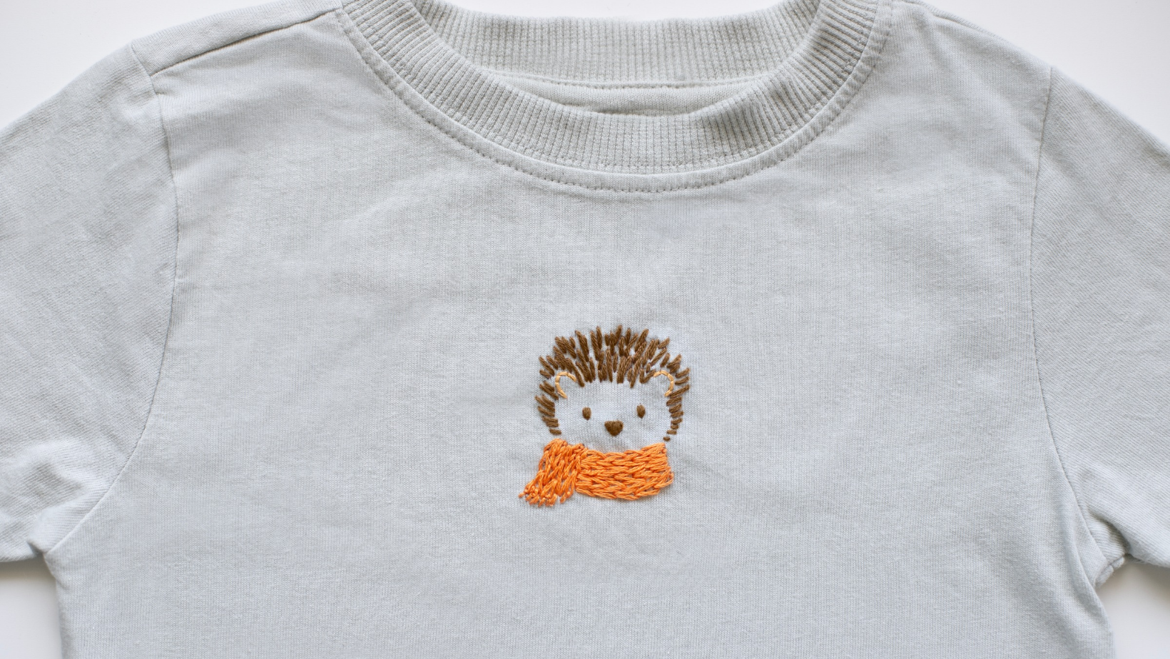 A hedgehog wearing an orange scarf, embroidered on the chest of a pale grey sweatshirt.
