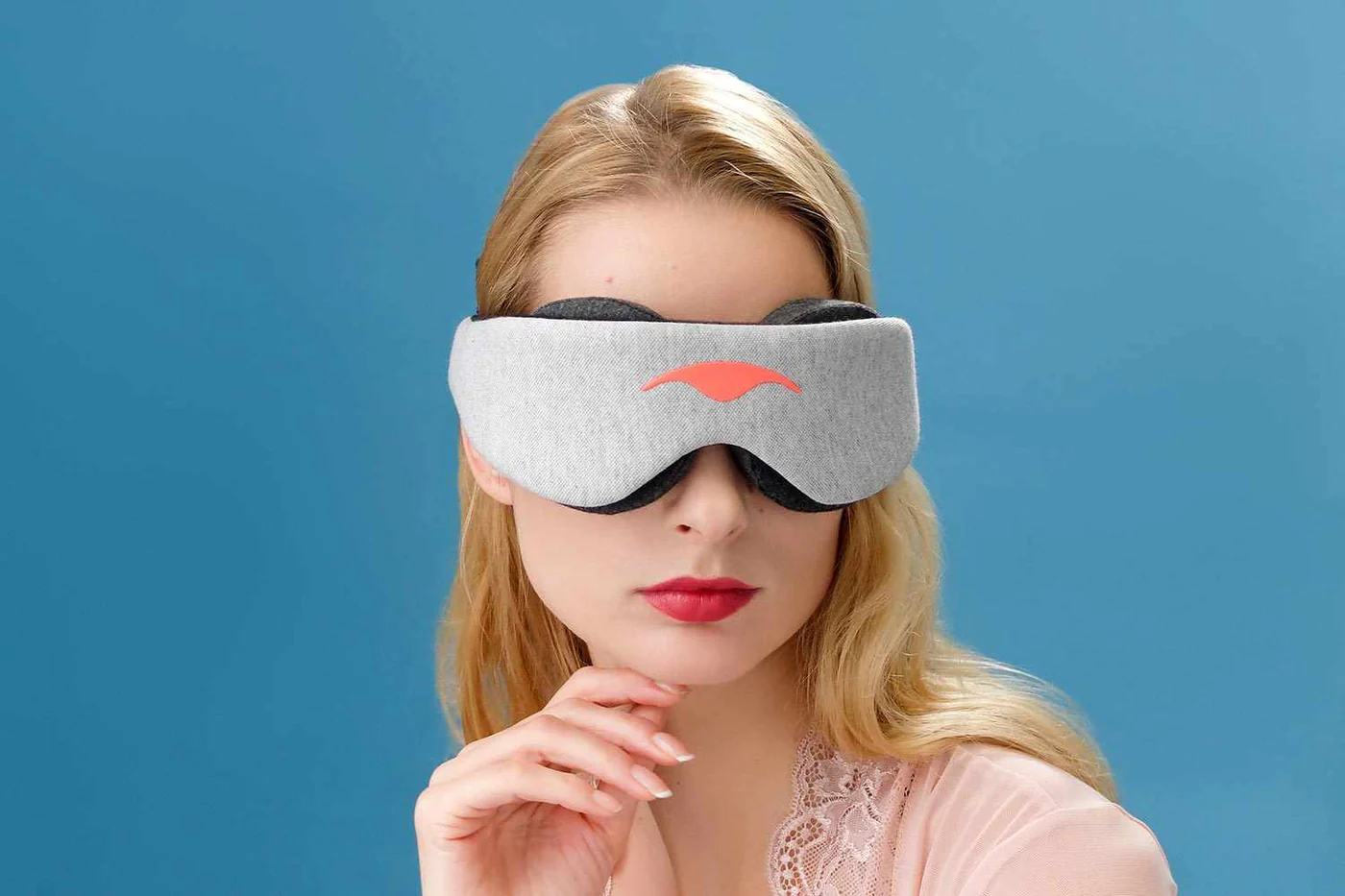A blonde girl holding the tip of her chin while wearing a gray sleep mask for napping.