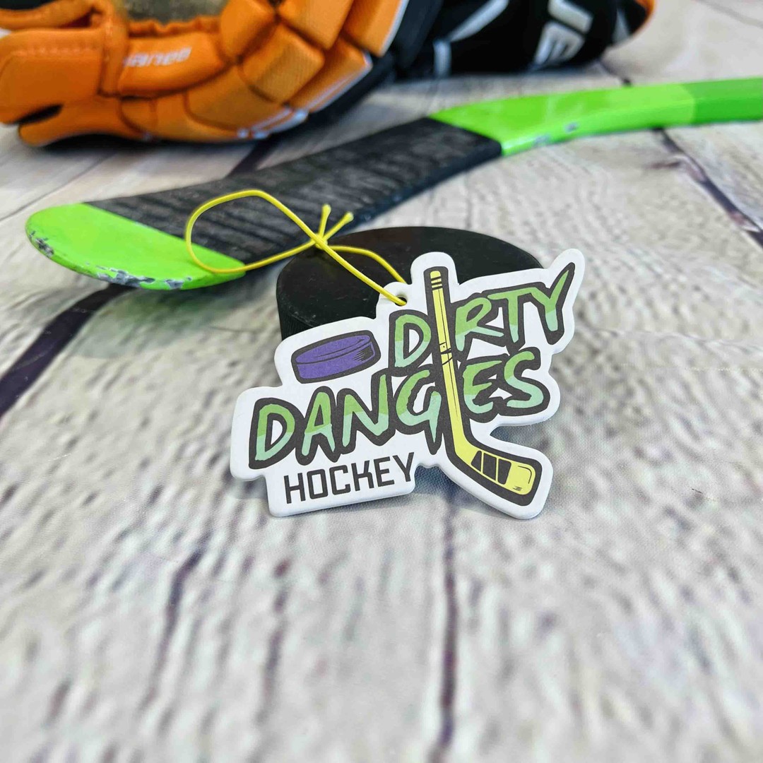A dirty dangles hockey car air freshener sits on a hockey puck with a hockey stick on a wood background. Dirty dangles hockey
