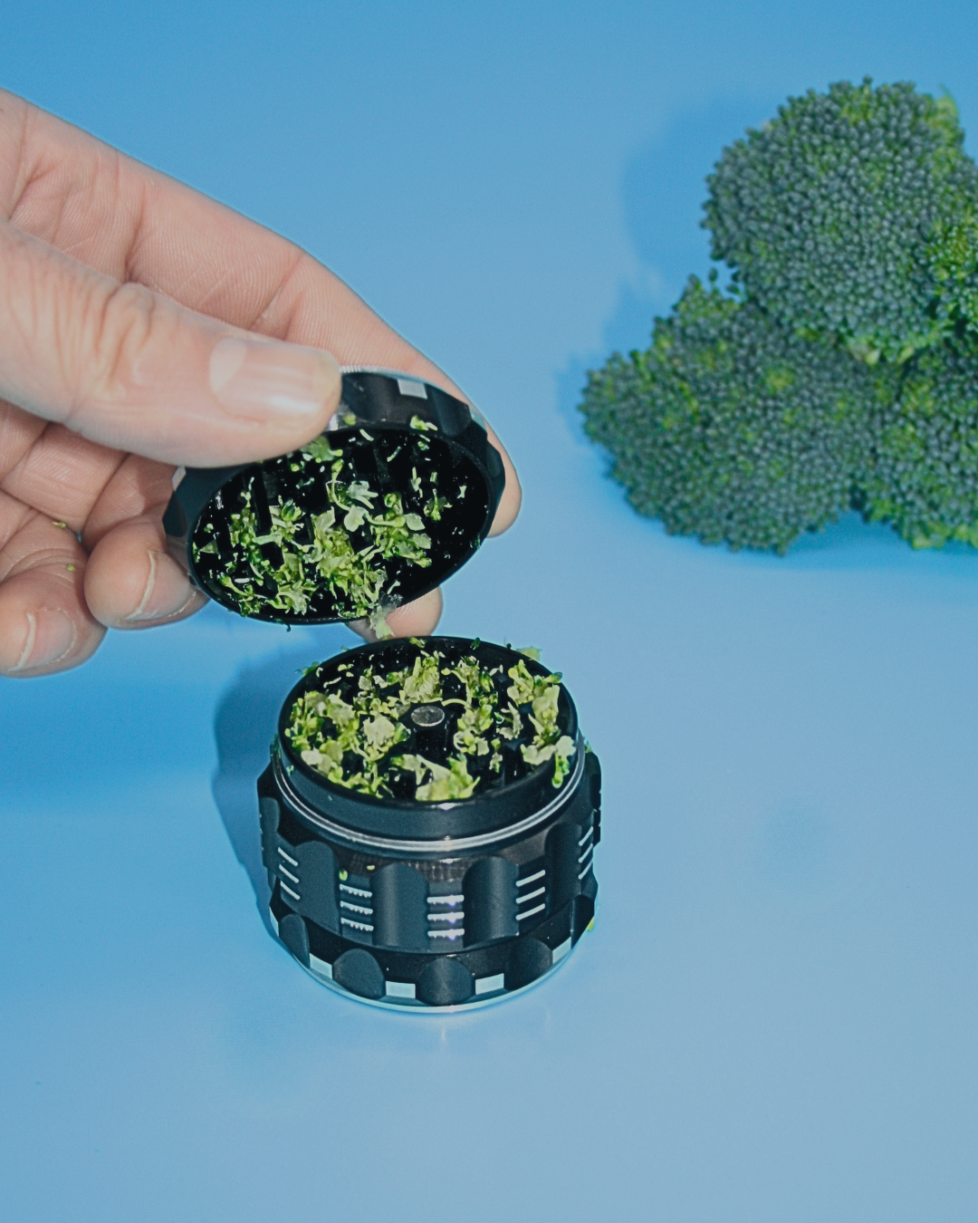 Close-up view of a hand loading material into the open diamond-cut grinding chamber of a black aerospace-grade aluminum Prism Herb Grinder, illustrating herb preparation.