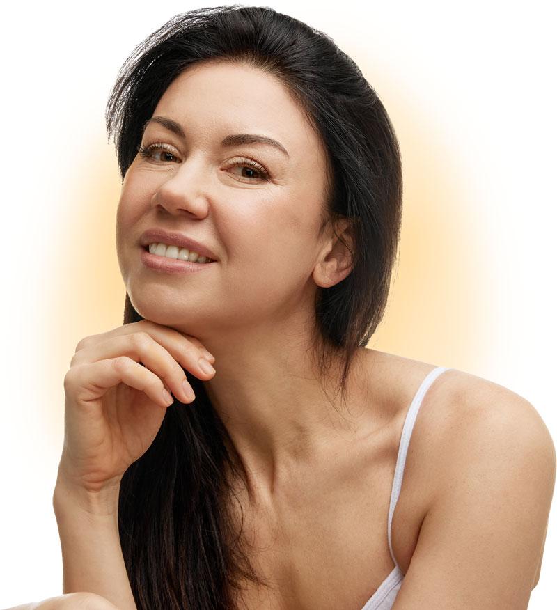 Smiling woman with clear, healthy skin and long dark hair, resting her chin on her hand, wearing a white strap top.