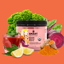 A jar of Revive Beets + Roots surrounded by 2 slices of lemon and a glass filled with red juice topped with green leaves on the left side. On the right side is a bunch of turmeric seated on top of a handful of a handful of orange powder. On the background is a sliced beet and a bunch of kale.