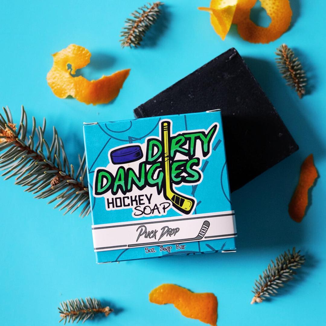 A bar of dirty dangles hockey soap puck drop scent on a blue background with pine and orange peel.