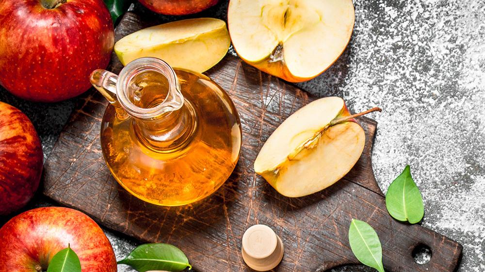 Grey background with wood cutting board, red apples with one cut in half, leaves and a glass bottle pitcher with apple cider vinegar.