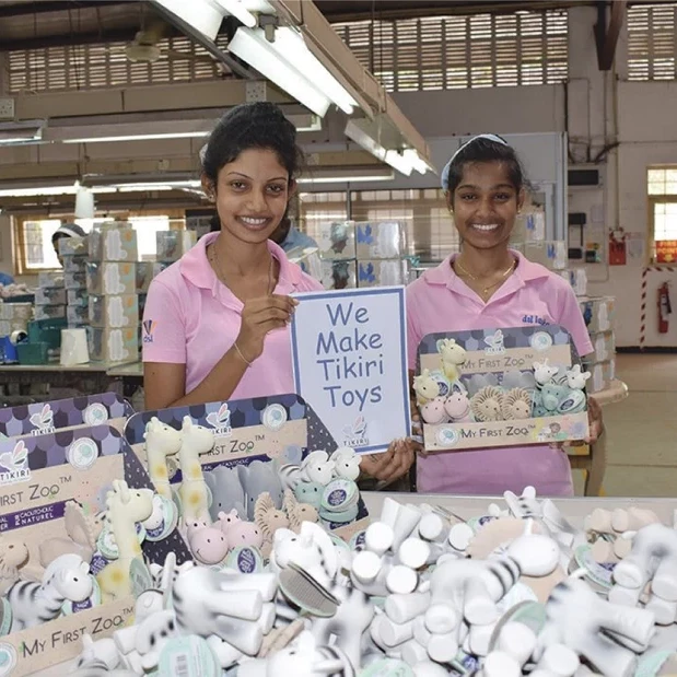 Two girls surrounded by baby toys holding up a sign saying We Make Tikiri Toys