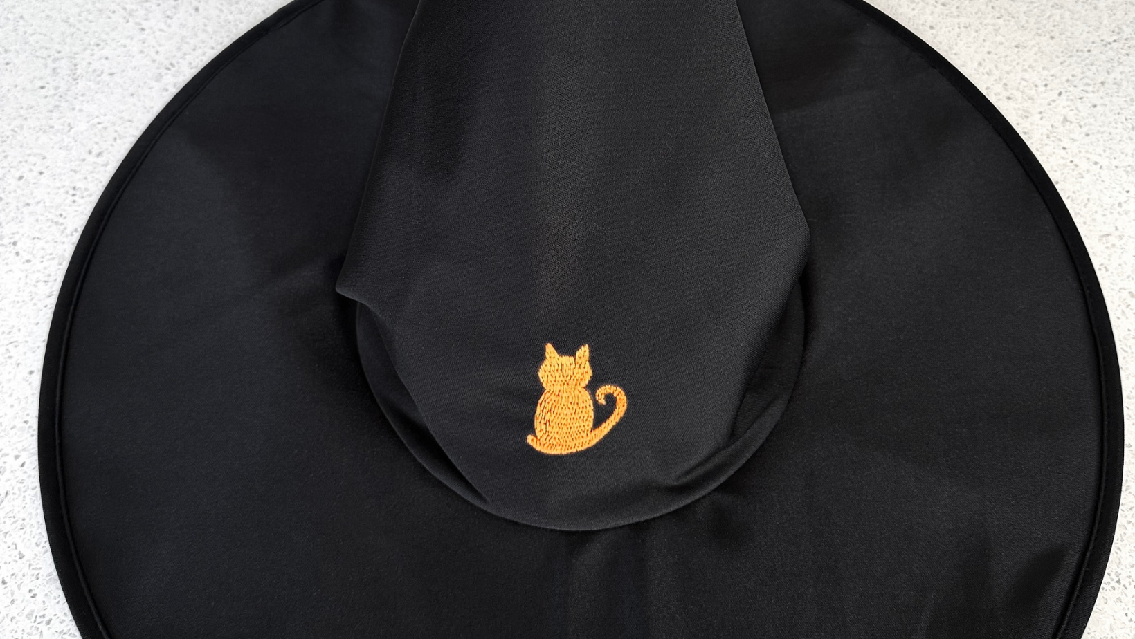 An orange cat embroidered onto the crown of a black witch's hat.
