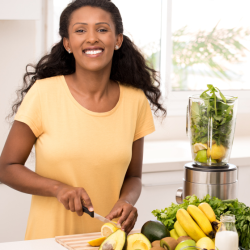 woman smiling at the camera as she cuts yellow and green fruits and veggies and puts them into a blender.