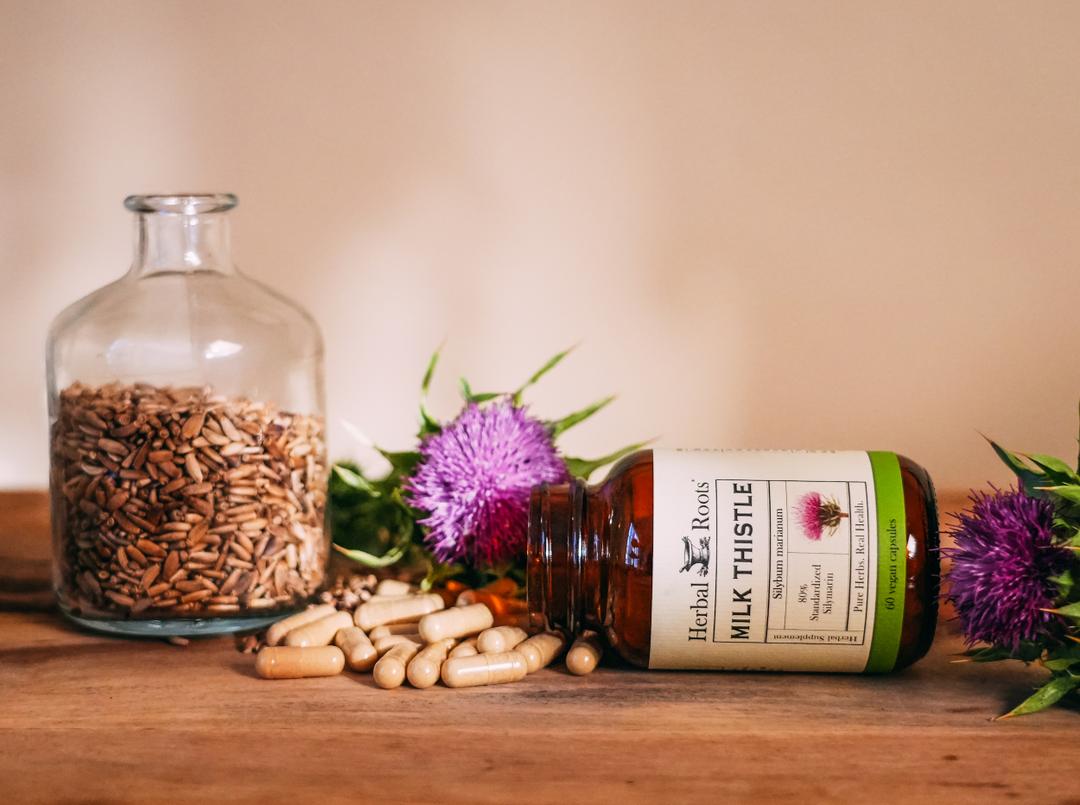 Herbal Roots bottle oh capsules spilling out next to a jar filled with milk thistle seed and two milk thistle flowers