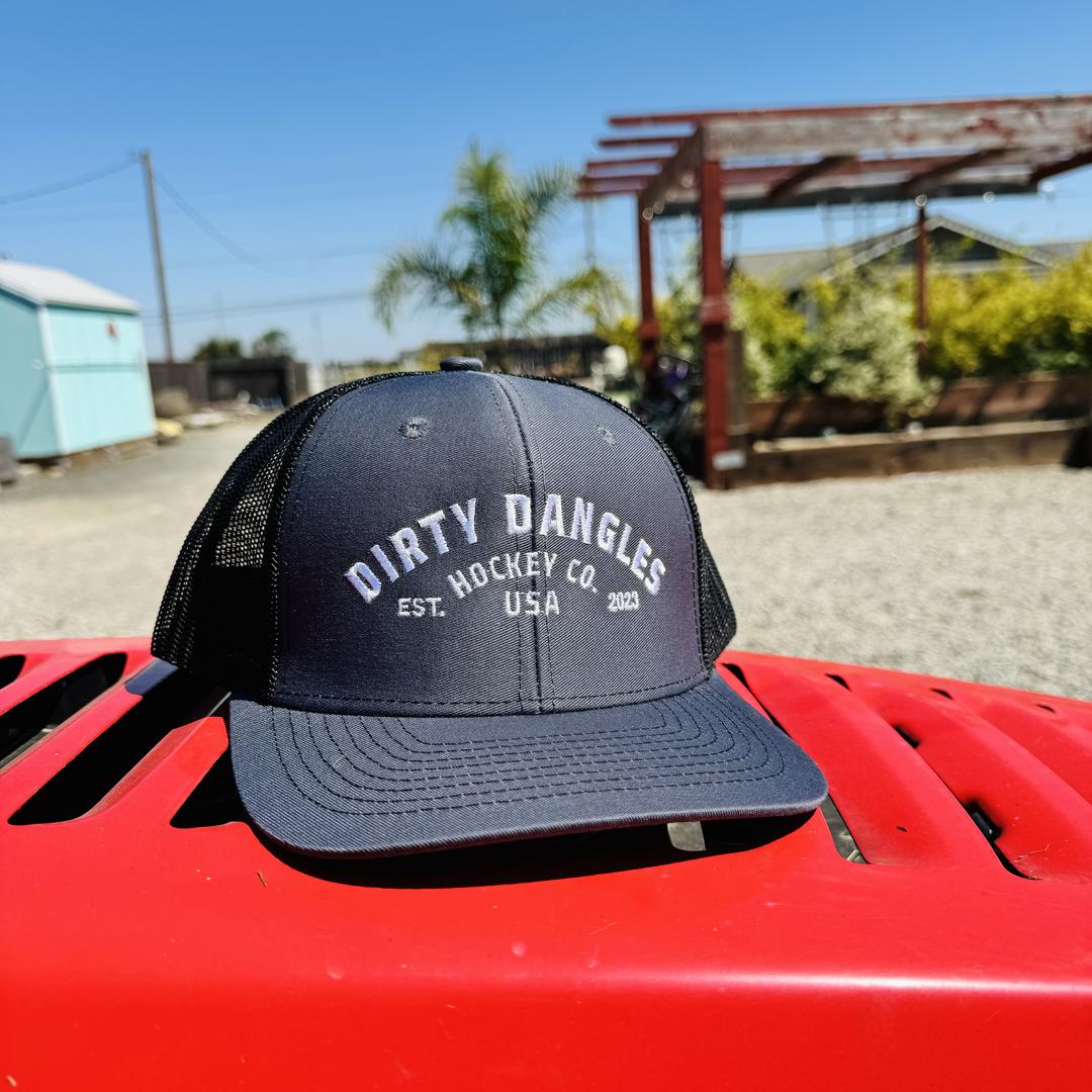 a dark gray and black dirty dangles hockey co mesh snapback trucker hat outside on a red lawn mower.