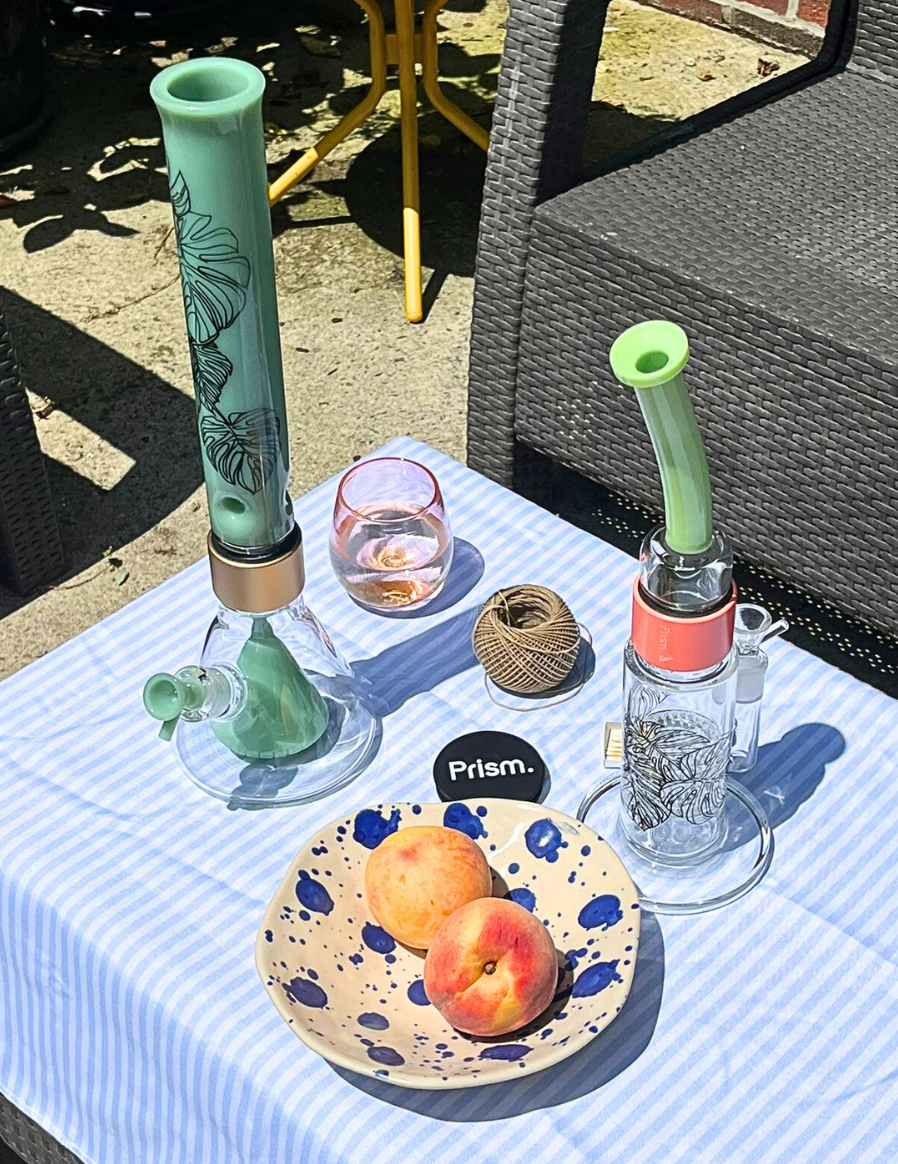 Summer tablescape featuring two Monstera Decal Prism bongs on a striped tablecloth: A Mint Green Percolated Beaker Bong and a Key Lime Green Honeycomb Bong. The scene, complete with peaches and a glass of wine, highlights the decals' fit into a relaxed, biophilic home decor and lifestyle setting.