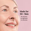 A smiling older Caucasian woman with blue eyes looking upward against a soft pink background. Text reads: Made for 50+ Skin — no dryness, no tightness, no added silicones.