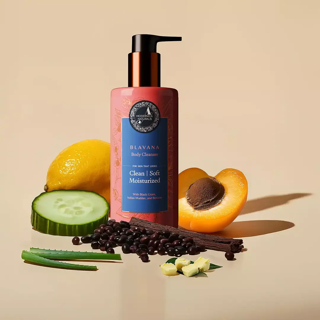 4. The image shows an arrangement of various natural ingredients around the blavana cleanser bottle. Ingredients include: A pink lotus flower Sliced lemons Whole lemons Cucumber slices Black gram manjistha sticks