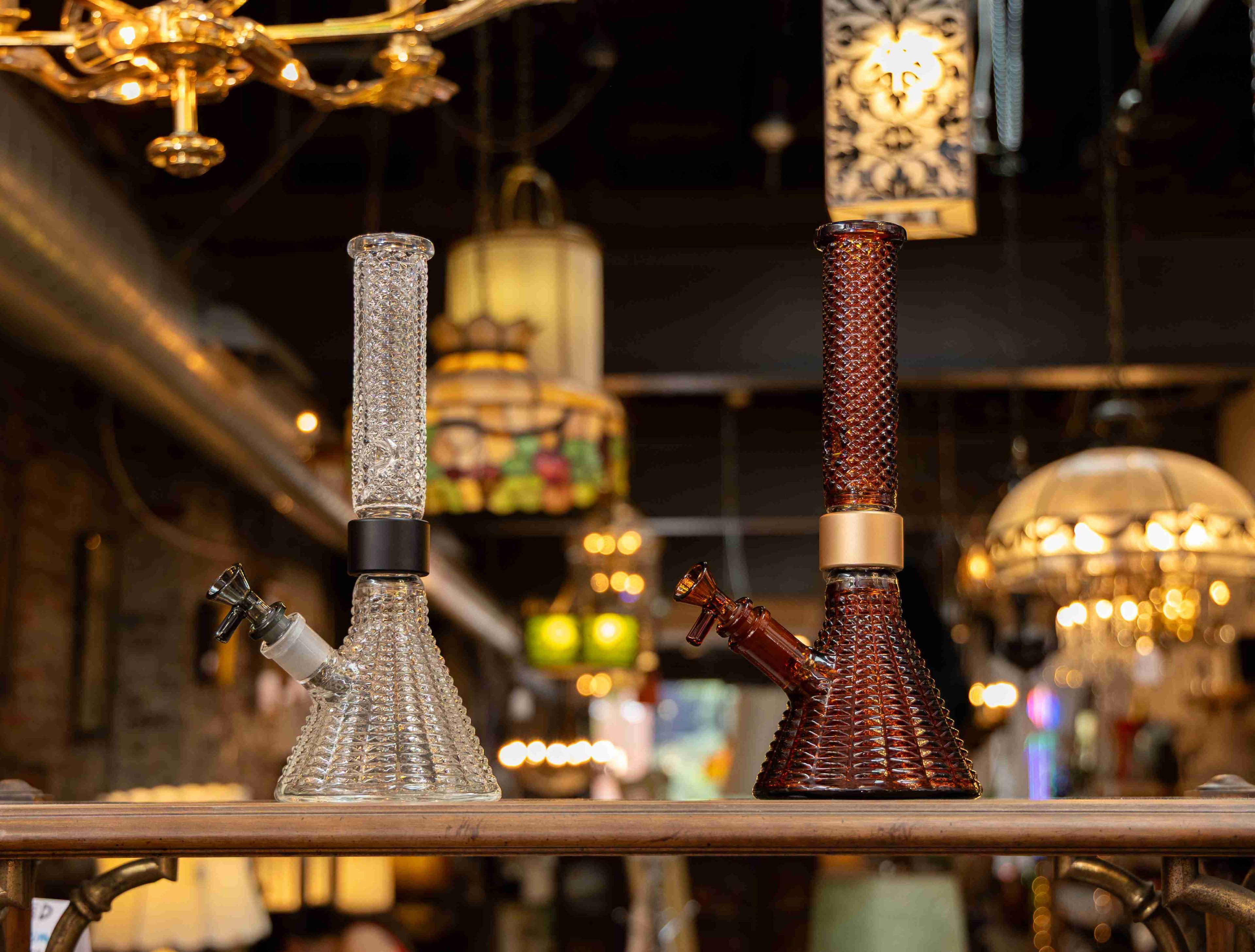 Side-by-side display of the limited-edition Prism x Jane West Twenties MINI Beaker Bongs, featuring the unique, tactile gridded diamond texture. The clear version (left) with black accents contrasts with the exclusive vintage amber version (right) with gold hardware. The bongs are photographed in a vintage Art Deco shop, reinforcing the 1920s elegance and centerpiece design.