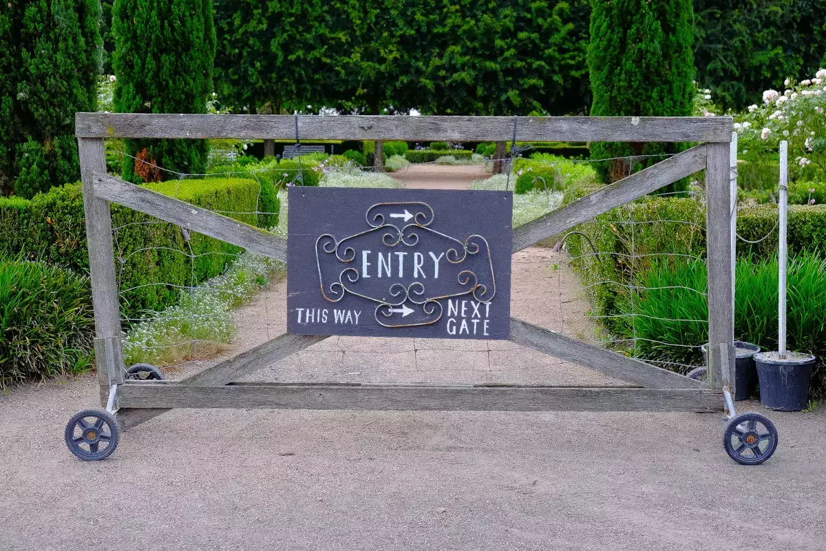 Rustic wooden garden gate with a decorative sign reading “Entry – This Way / Next Gate,” set along a gravel path bordered by manicured hedges and formal garden beds.