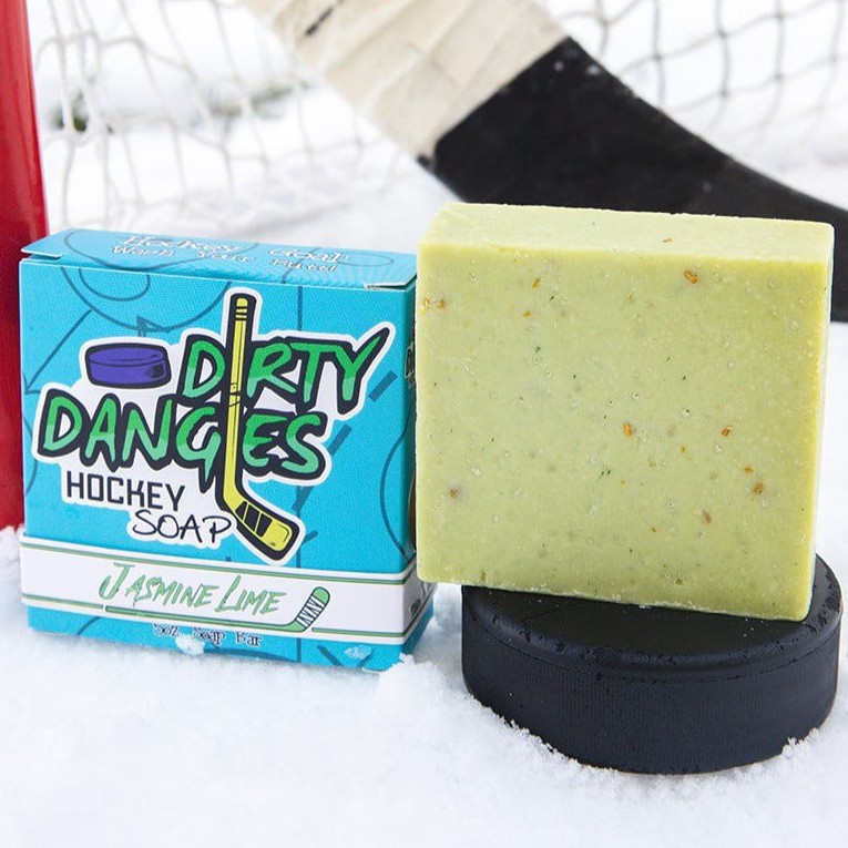 A green bar of dirty dangles hockey soap sits on a hockey puck in the snow with a hockey stick and a hockey goal. Jasmine lime scent