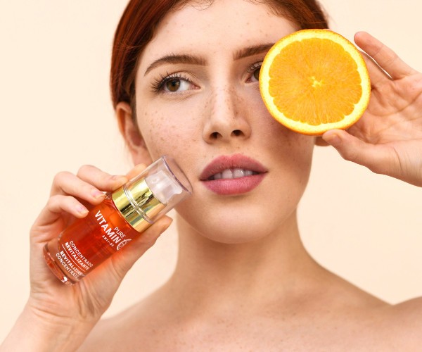 young woman with red hair holding a sliced orange in front of her eye and a bottle of Noche's Vitamin C Serum in front of her cheek