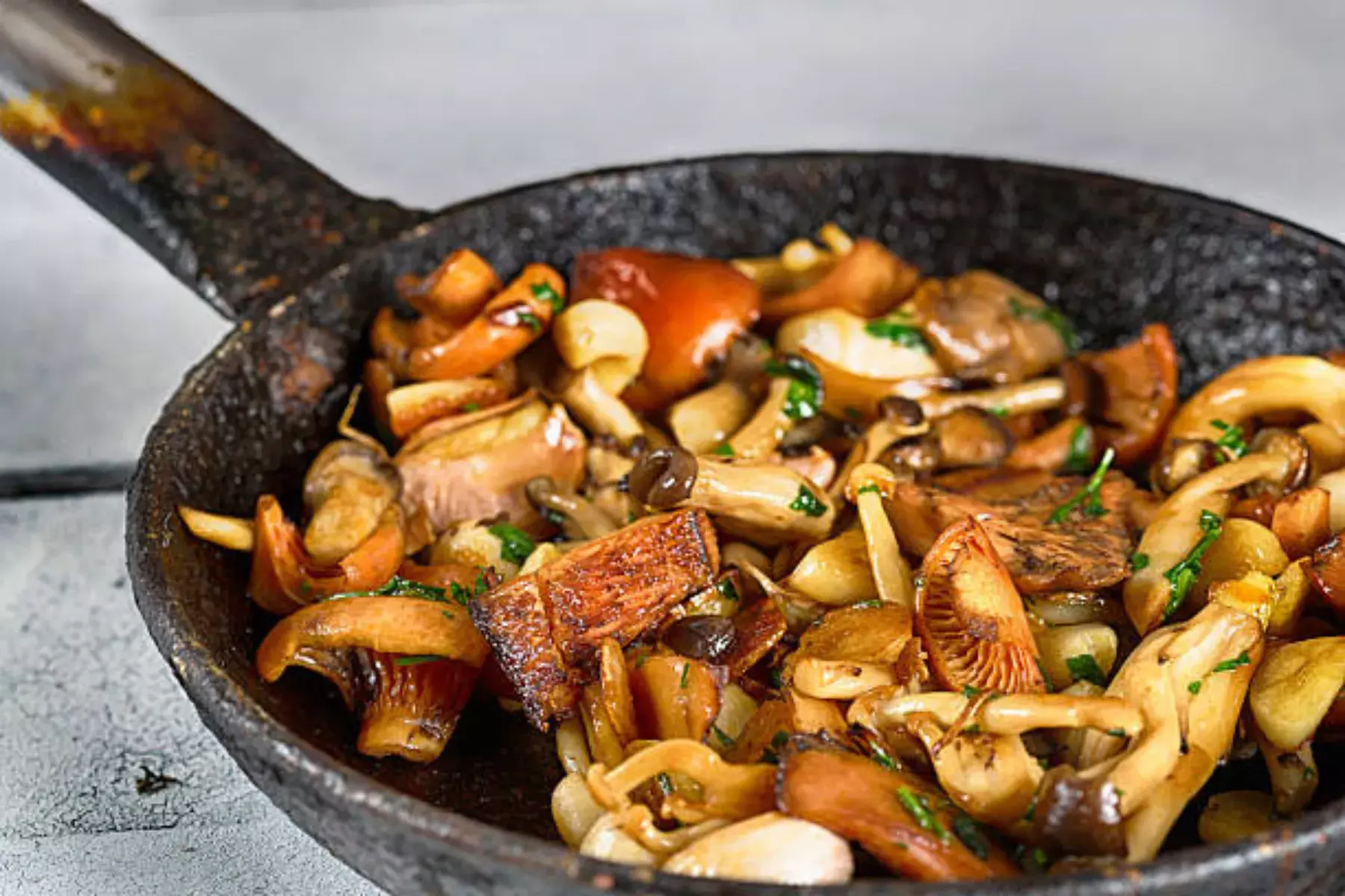 Sautéed mixed mushrooms in cast iron skillet - golden brown caramelized fungi medley including shiitake, cremini, oyster mushrooms with fresh parsley garnish, pan-seared cooking method, non-stick speckled cookware, culinary preparation, restaurant-style presentation, umami-rich dish, vegetarian side dish, high-heat cooking technique