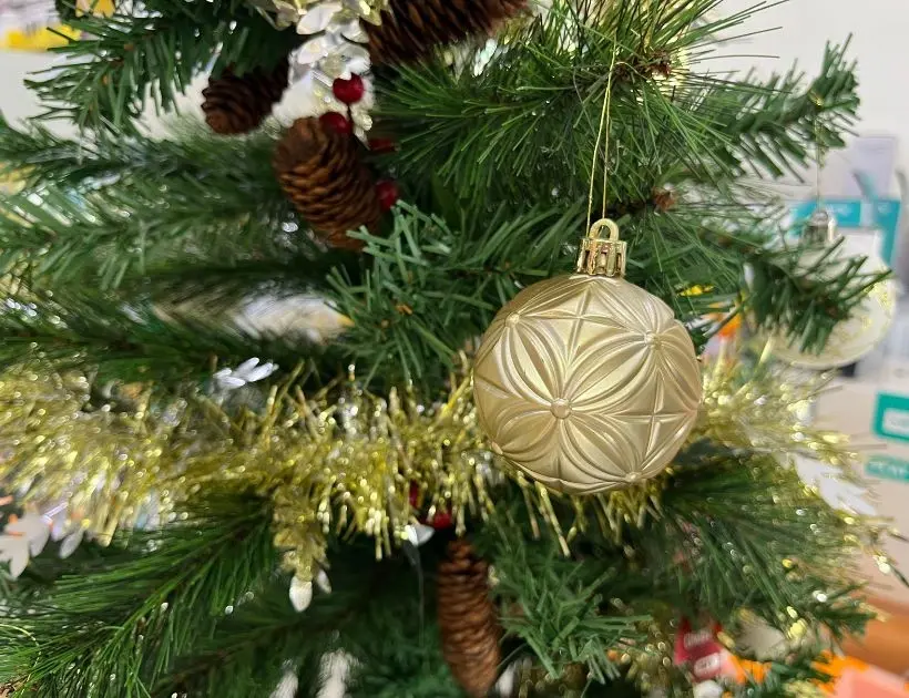 A gold Christmas ornament hanging on a decorated Christmas tree, surrounded by green branches and twinkling lights.