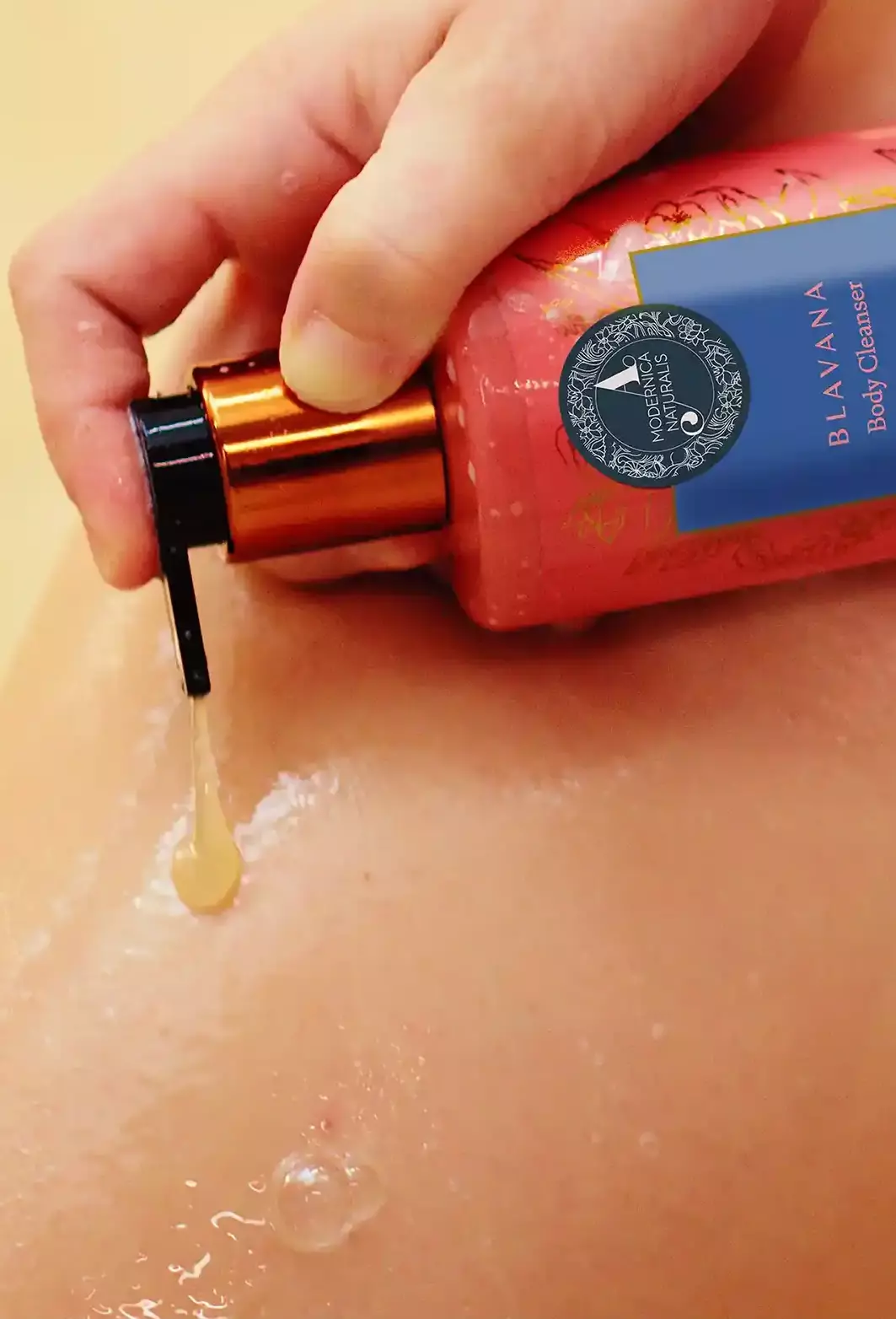 2. 1. The image shows a woman hand holding a Blavana cleanser bottle. She is gently squeezing the bottle, causing a clear liquid to come out of the bottle. The background is a neutral, beige color.